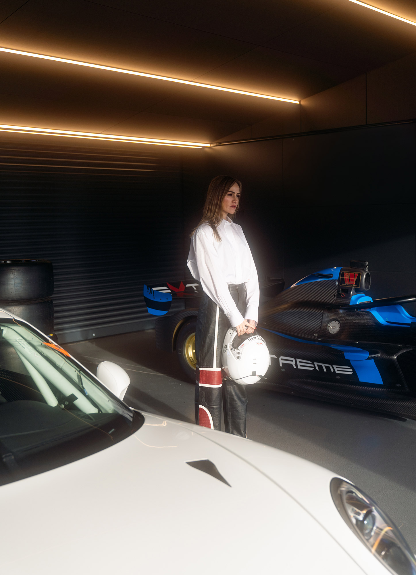 Carmen in a garage with two supercars holding a racing helmet