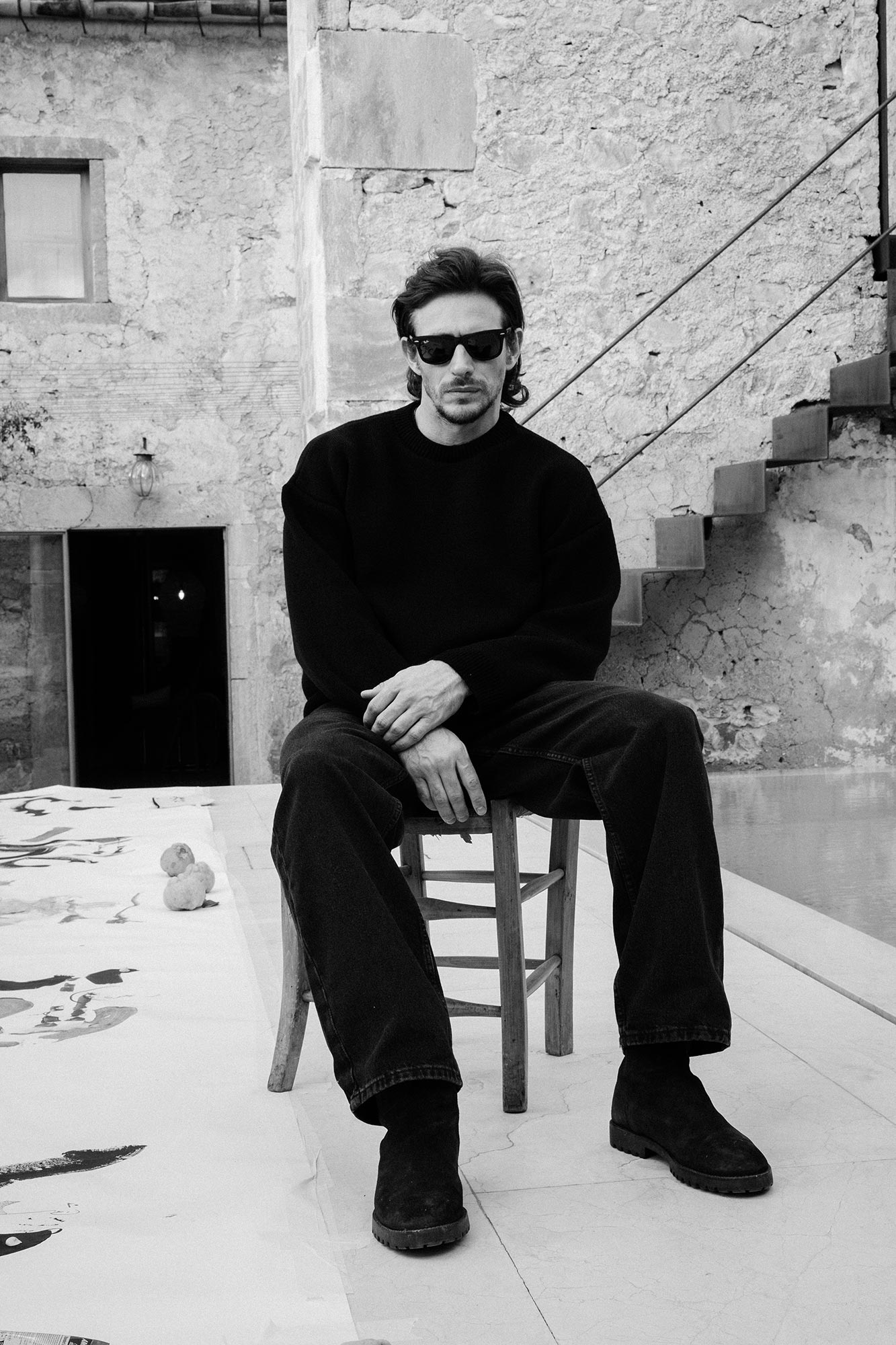 Jake sitting on a stool in all black clothing wearing black sunglasses