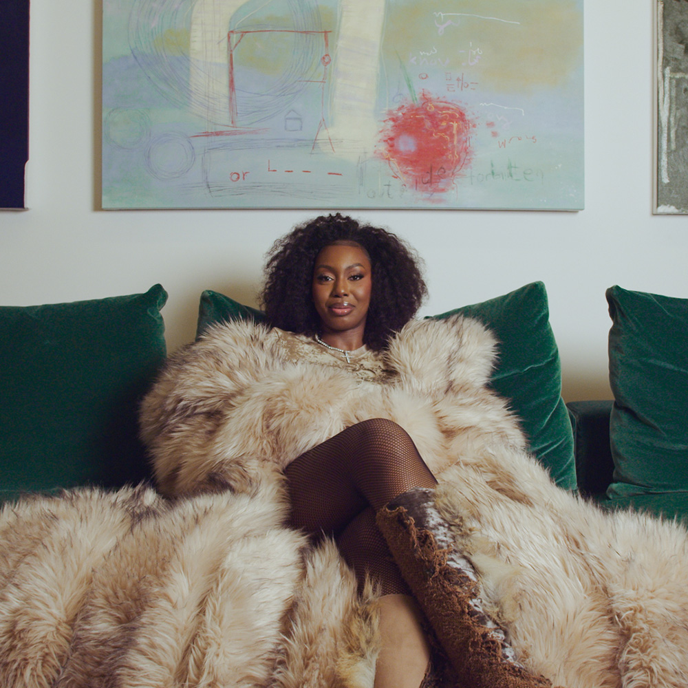 Ms Banks wearing a fur coat sat on a green sofa